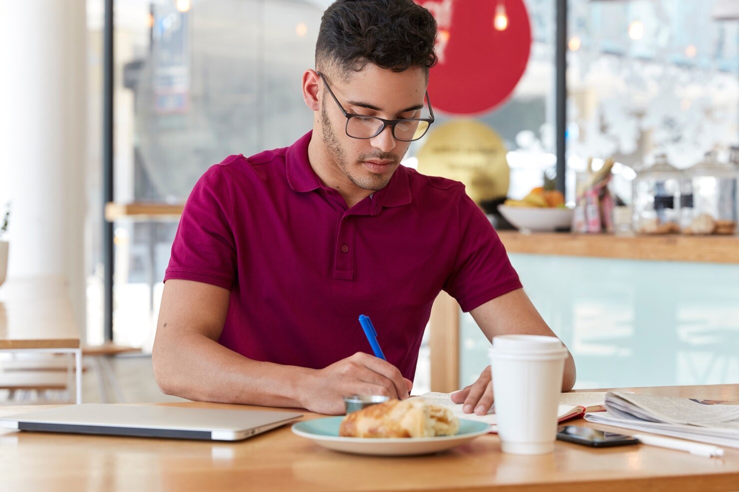 A young adult writes in a notebook beside a laptop, coffee, and breakfast at a cafe table.
