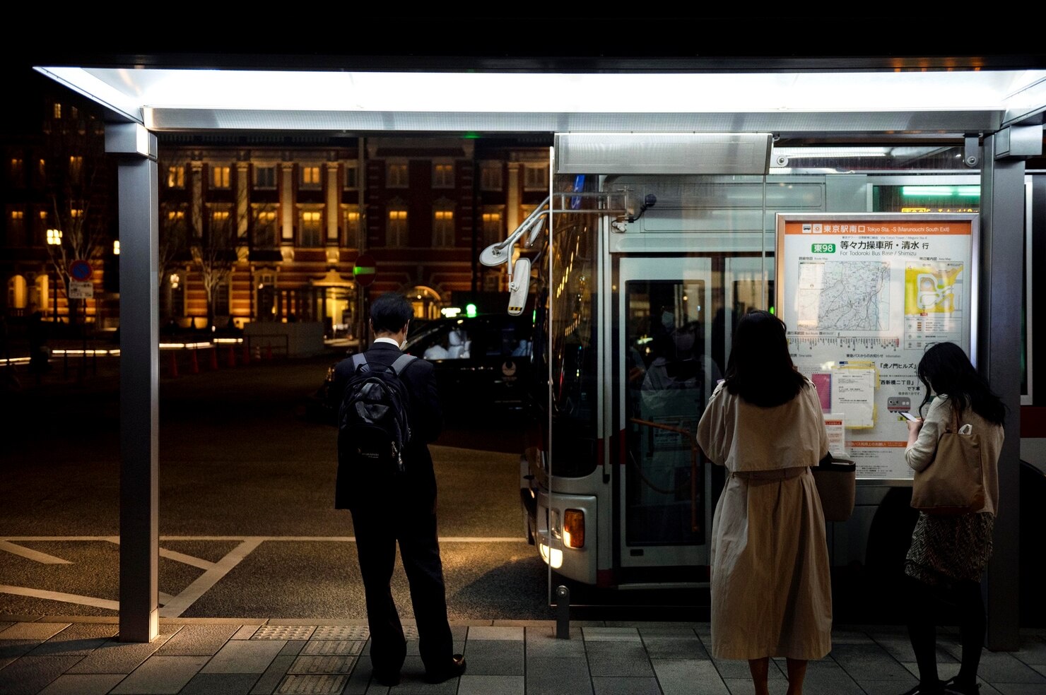 Three commuters wait beneath the bright lights of a bus shelter while the city around them is still dark.