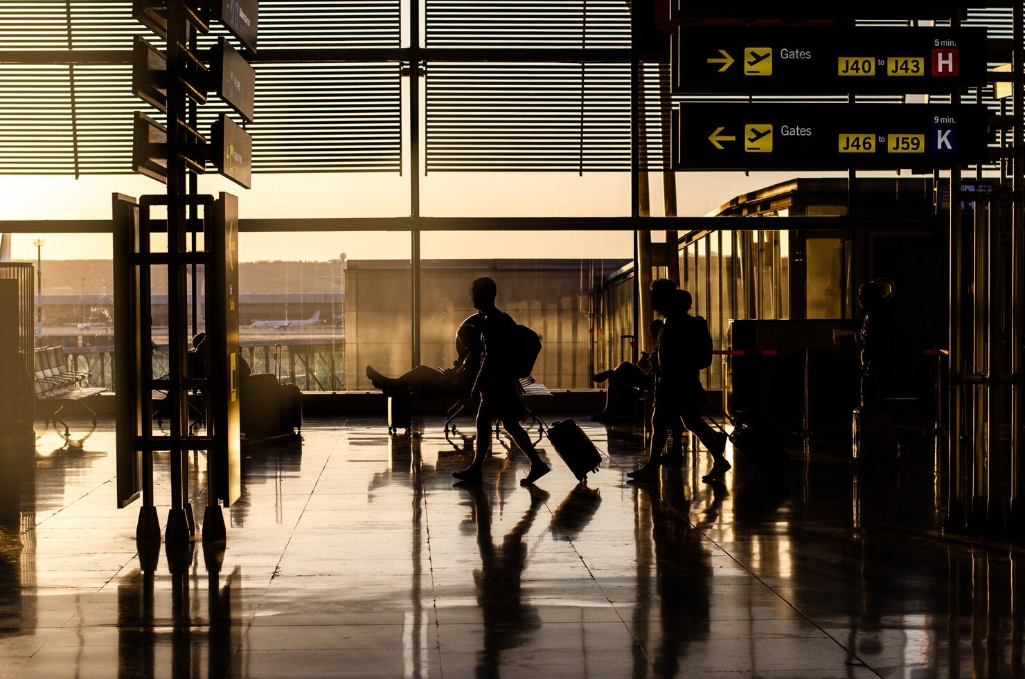 Silhouetted travelers walking through an airport terminal with rolling suitcases, illuminated by warm sunlight streaming through large windows. Overhead signs indicate directions to gates J40 to J43 and J46 to J59, with estimated walking times of 5 and 9 minutes. The polished floor reflects the figures and light, creating a dynamic and anticipatory atmosphere typical of travel.