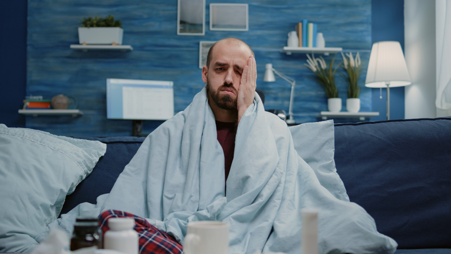 Man sitting on a dark blue sofa wrapped in a light blue blanket, wearing red plaid pajama pants. He is surrounded by various medicine bottles, a mug, and a tissue box on the table in front of him, suggesting he is unwell. The background features a modern living room with blue walls, shelves with books and plants, a desk lamp, and a computer monitor. The atmosphere feels quiet and subdued, conveying a sense of illness and rest at home.