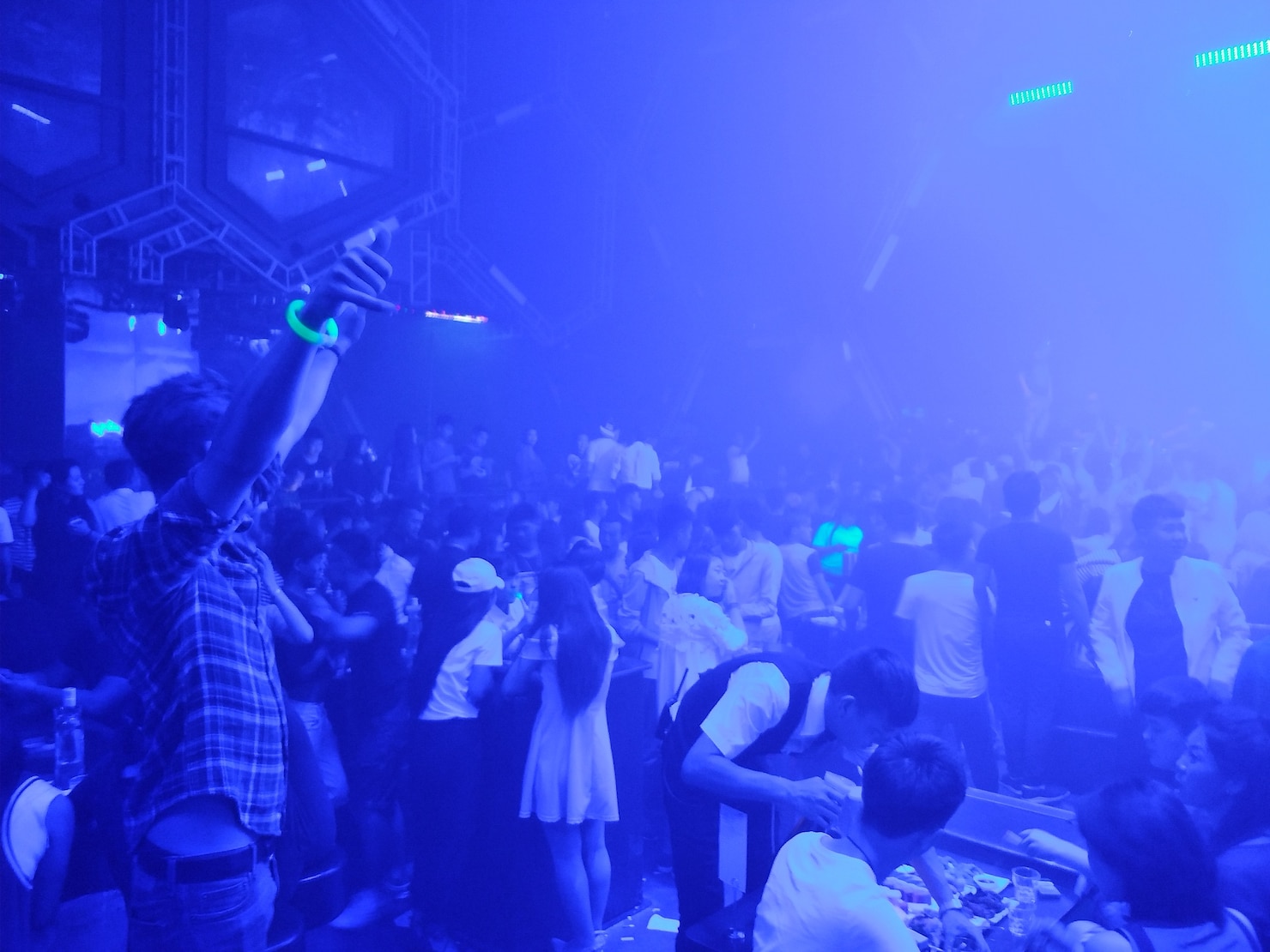 Crowded nightclub or party scene with people standing, dancing, and socializing under blue lighting. A person in the foreground raises an arm with a glowing bracelet, while others gather around tables and the bar. The atmosphere is energetic and lively, with modern geometric decor and bright LED lights overhead.