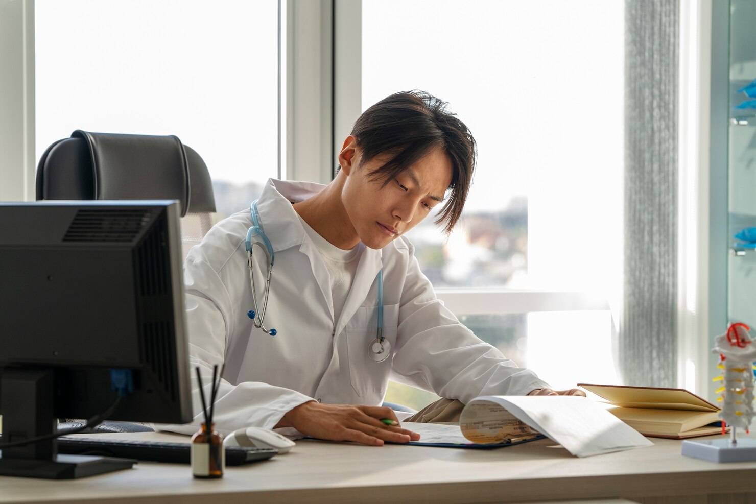 Doctor wearing a white lab coat and stethoscope sits at a desk in a bright office, reviewing documents and writing notes. The workspace includes a computer monitor, books, and medical supplies, with large windows letting in natural light. The environment feels professional and focused.