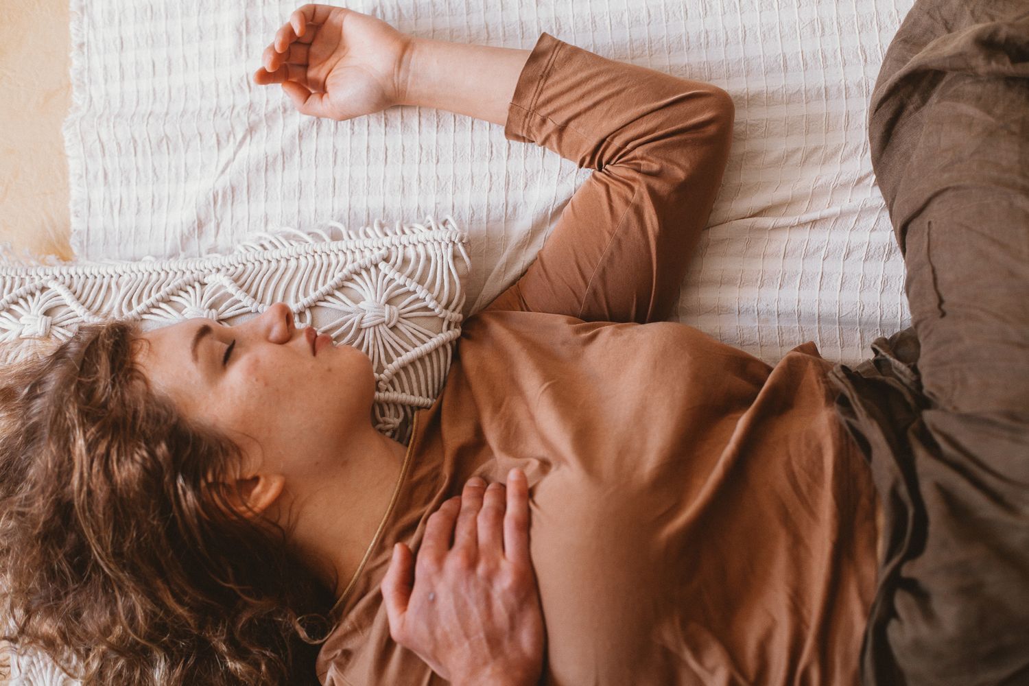 A woman sleeps across a bed in brown clothing, resting with one arm above her head.