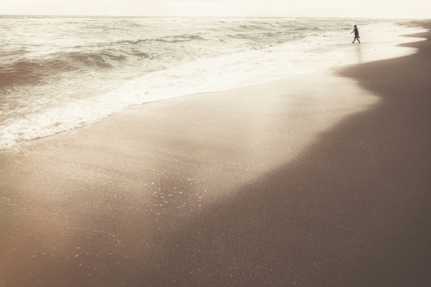A person walks along a quiet, sunlit beach, reflecting the outdoor light and movement that can make sleep feel easier on vacation.