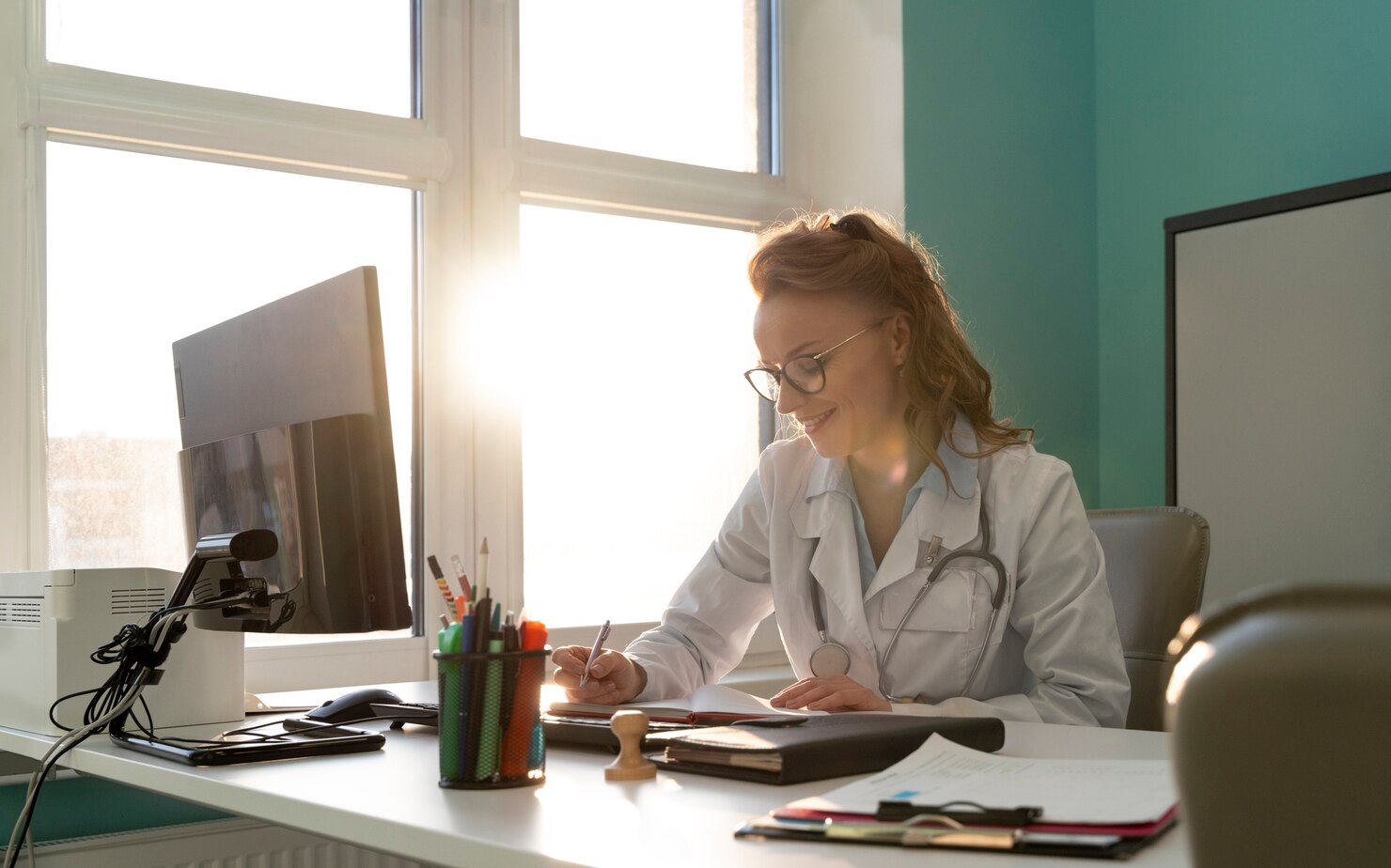 Doctor in a white lab coat and stethoscope sits at a desk, writing notes on paper while reviewing documents. The workspace features a computer monitor, books, and medical supplies, with large windows allowing sunlight to fill the bright office. The environment feels professional, organized, and focused.