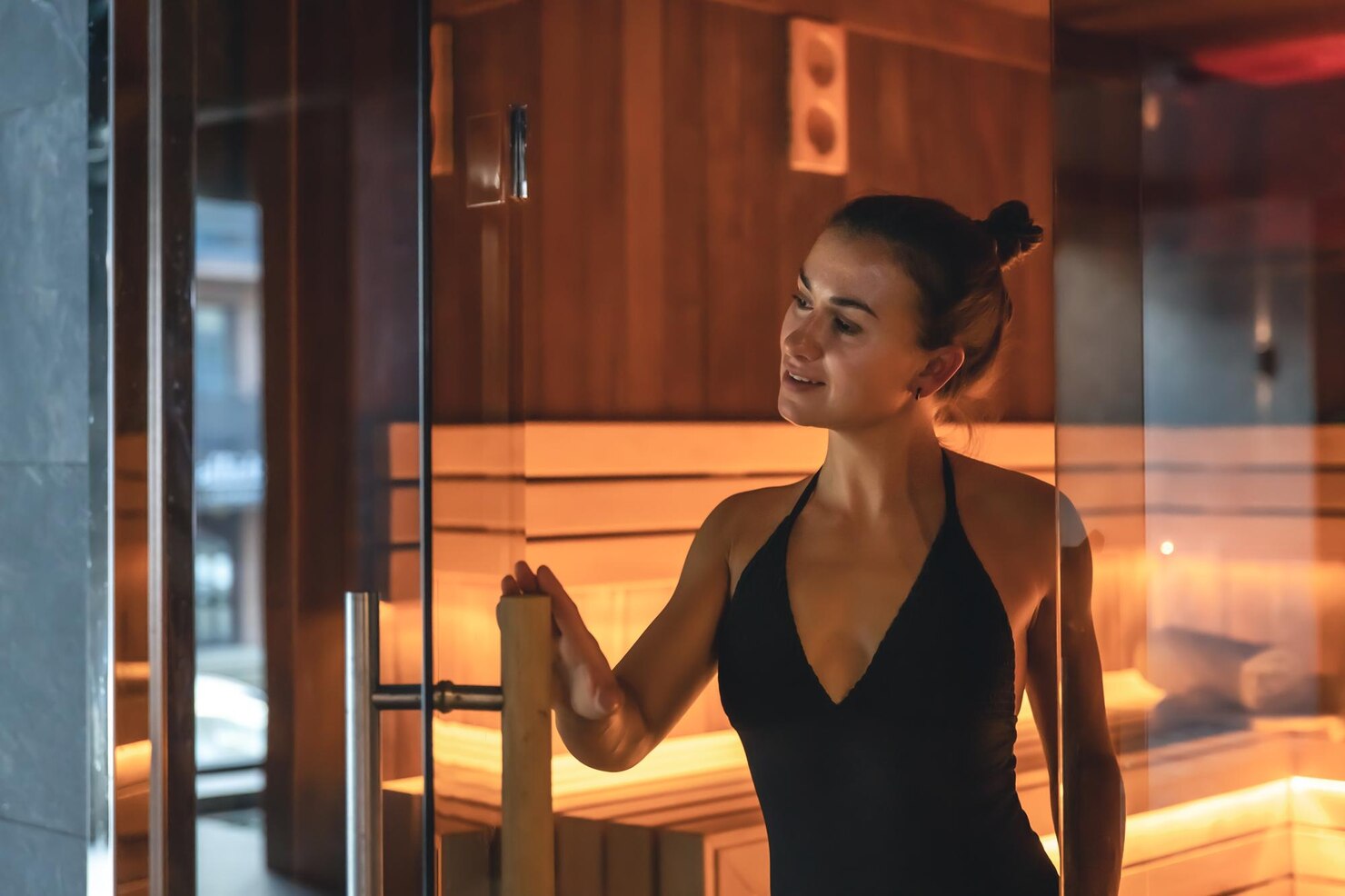 Woman wearing a black halter swimsuit stands at the entrance of a wooden sauna, holding the glass door open with one hand. The sauna interior is warmly lit with wooden benches and paneling, creating a cozy and relaxing atmosphere. The overall mood is calm and tranquil.
