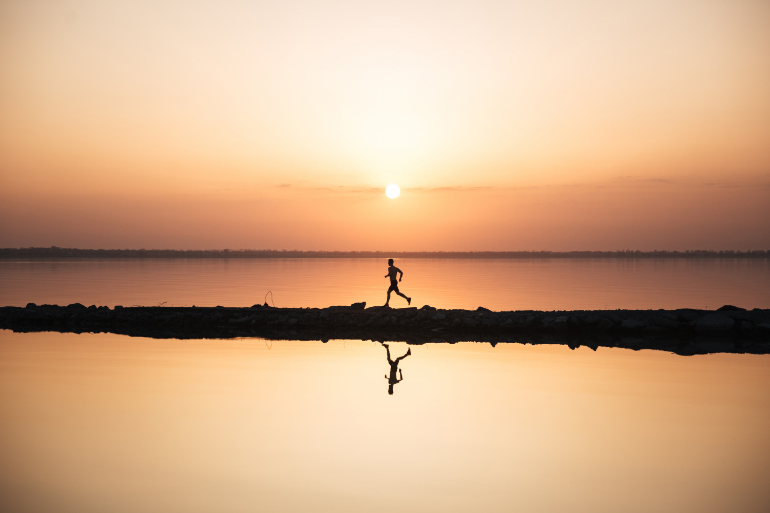 Silhouette of a person running along a narrow path beside a calm body of water at sunrise or sunset. The sun is low on the horizon, casting a warm orange glow across the sky and reflecting on the still water below. The runner's reflection is visible in the water, and the overall scene feels peaceful, serene, and energizing.