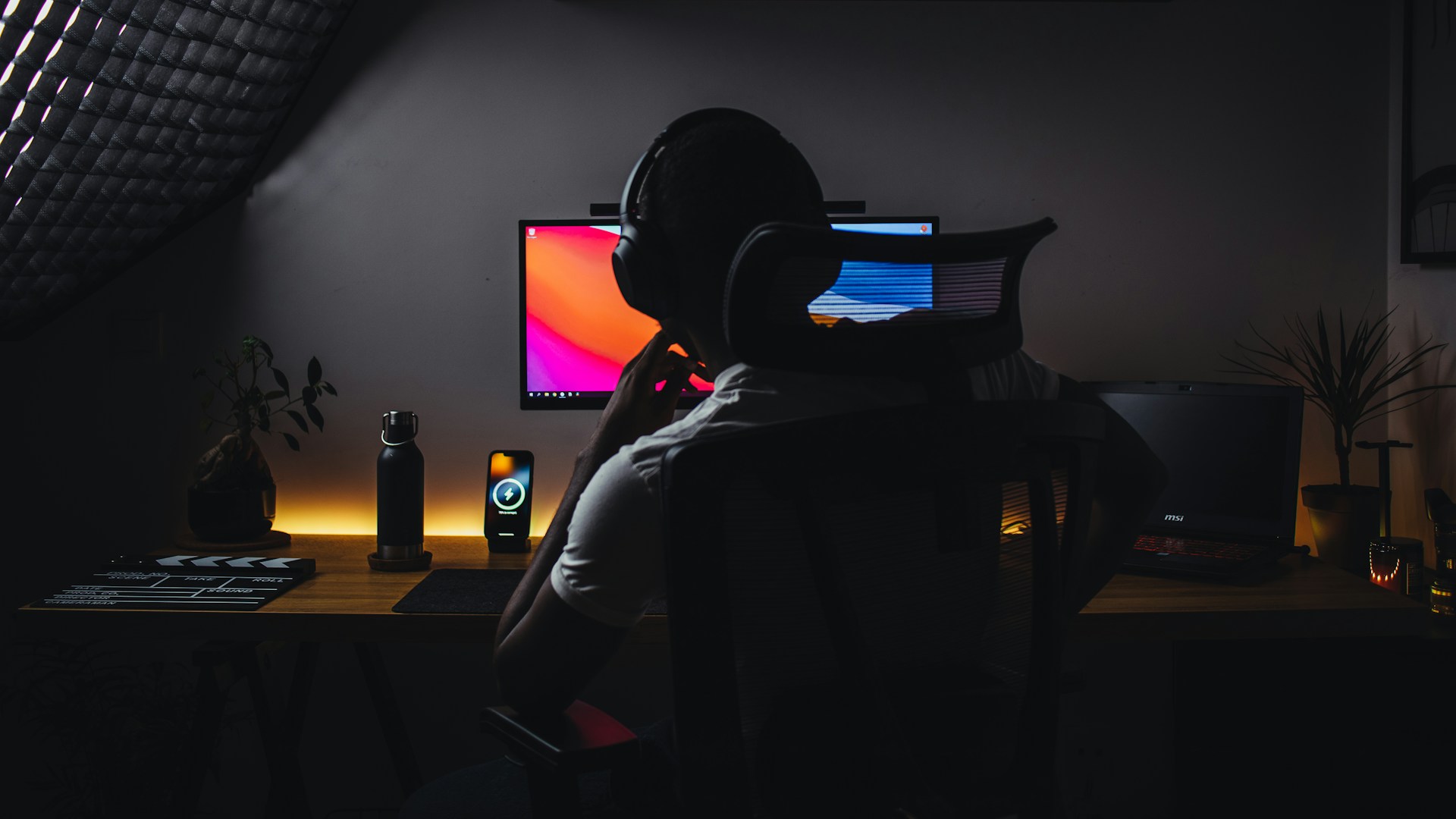 A person sits in a dark room at a desk, wearing headphones and facing a dual-monitor computer setup with colorful screens. The workspace is illuminated by soft ambient lighting and includes a water bottle, smartphone on a wireless charger, laptop, plants, and various desk accessories. The environment feels modern, focused, and slightly moody, suggesting late-night work or gaming.