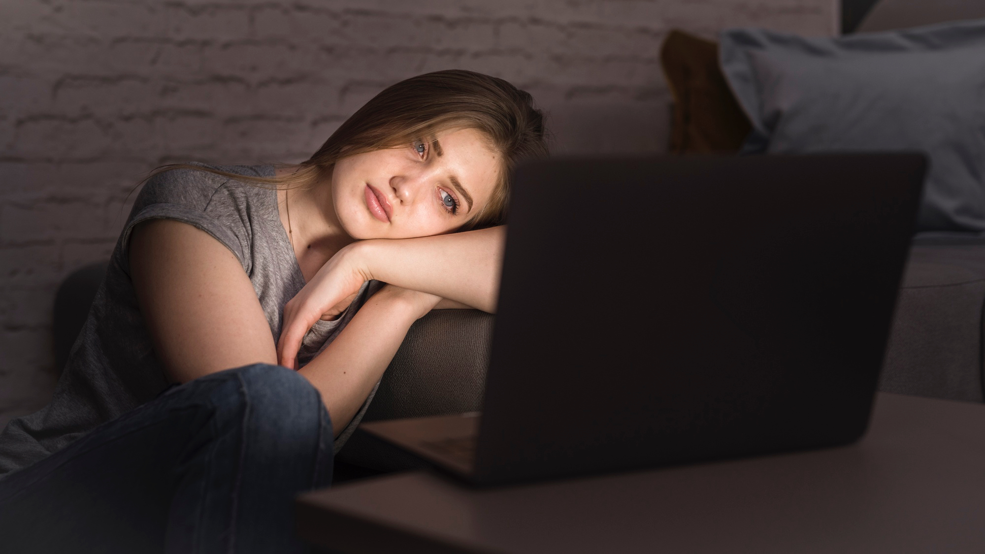A person sits cross-legged on a bed at night, using a laptop that casts a blue glow on their face. The room is dark, with white sheets and a pillow, and the mood is focused and slightly tense, suggesting late-night work or study.