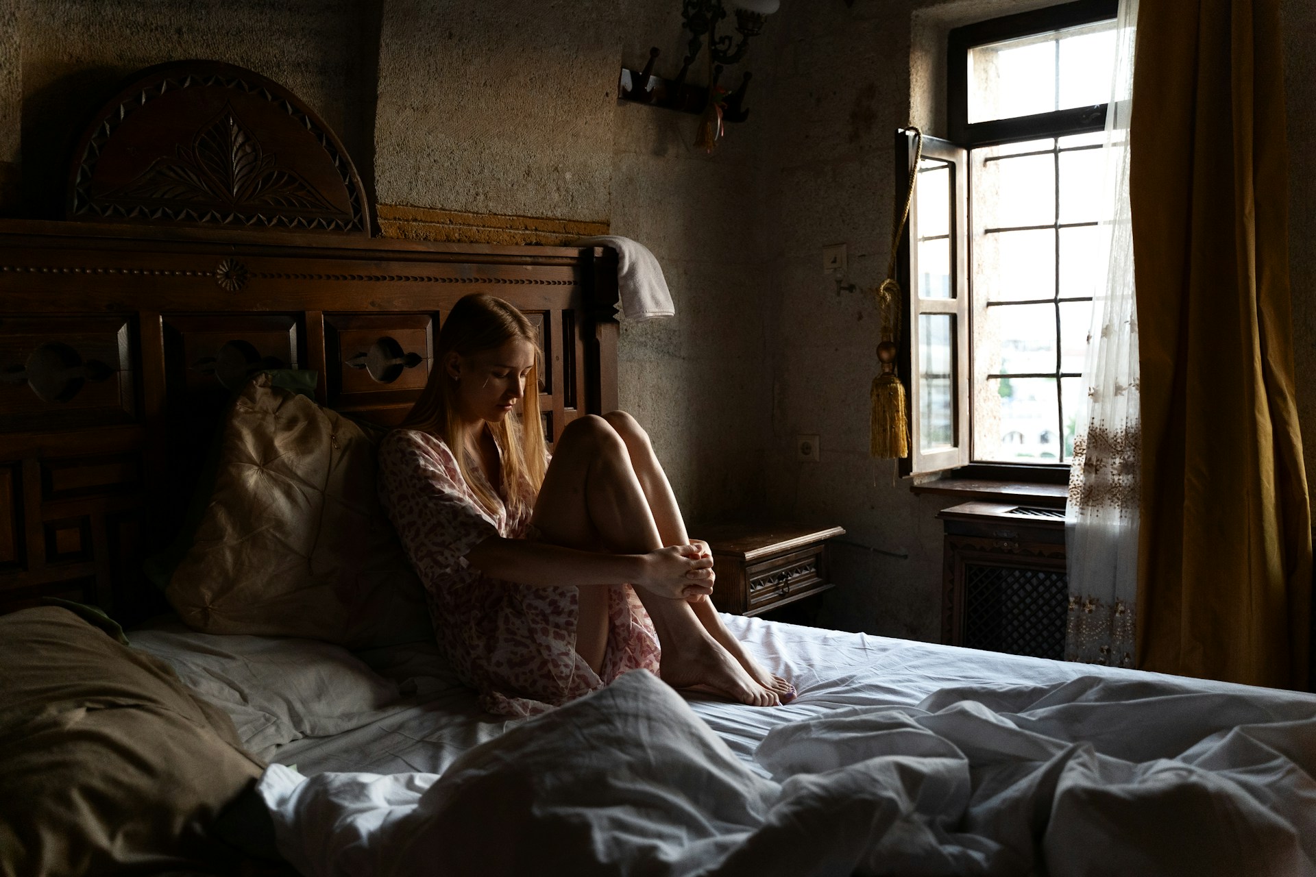 A woman sits on the edge of a bed in a softly lit bedroom, her head resting in her hand and her posture slouched. The bed has white sheets and a pillow, and the environment feels quiet and somber, suggesting fatigue or poor sleep quality.