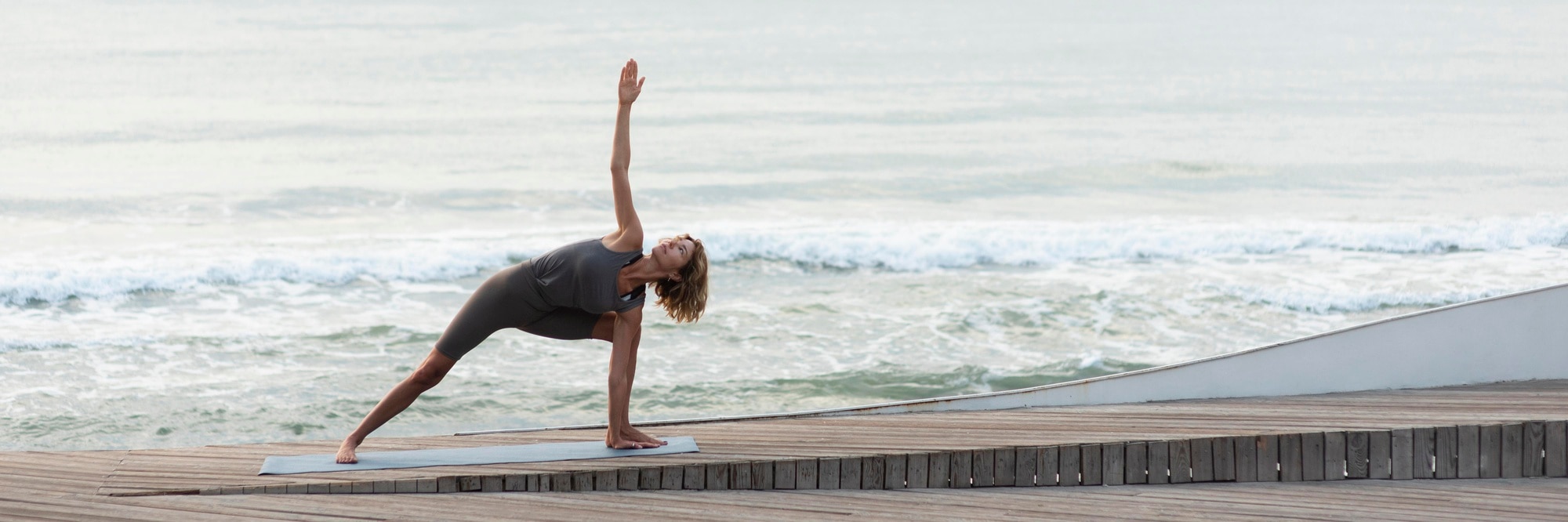 A woman in a gray athletic outfit practices a yoga pose on a mat outdoors on a wooden deck by the ocean. She is in a deep lunge with one hand touching the mat and the other arm extended upward, looking toward the raised hand. Gentle waves and a calm sea are visible in the background, creating a peaceful and focused atmosphere.