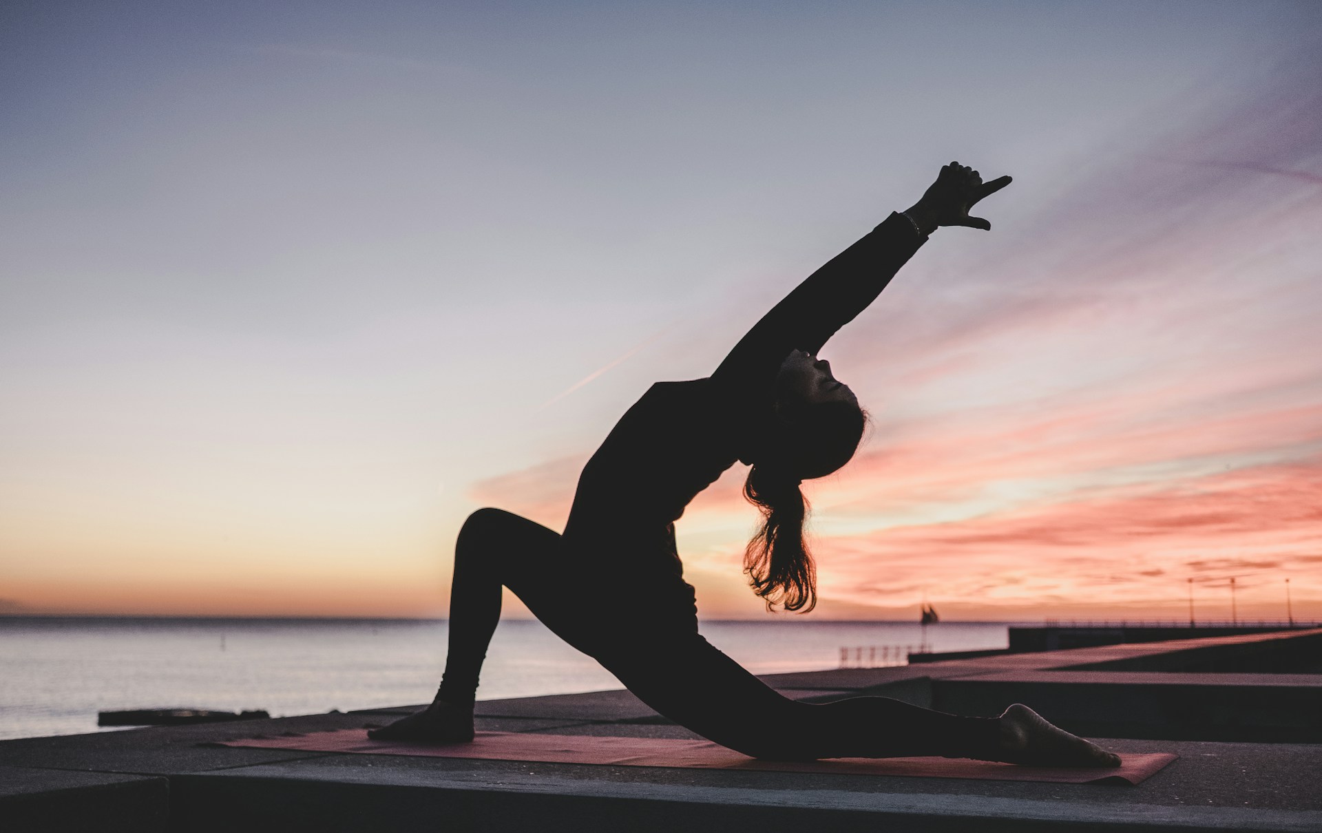 A person in athletic clothing performs a yoga stretch on a mat outdoors at sunset, with one knee bent forward and arms extended overhead. The background features a calm ocean and a colorful sky with soft clouds. The atmosphere feels peaceful and focused, suggesting relaxation and mindfulness.