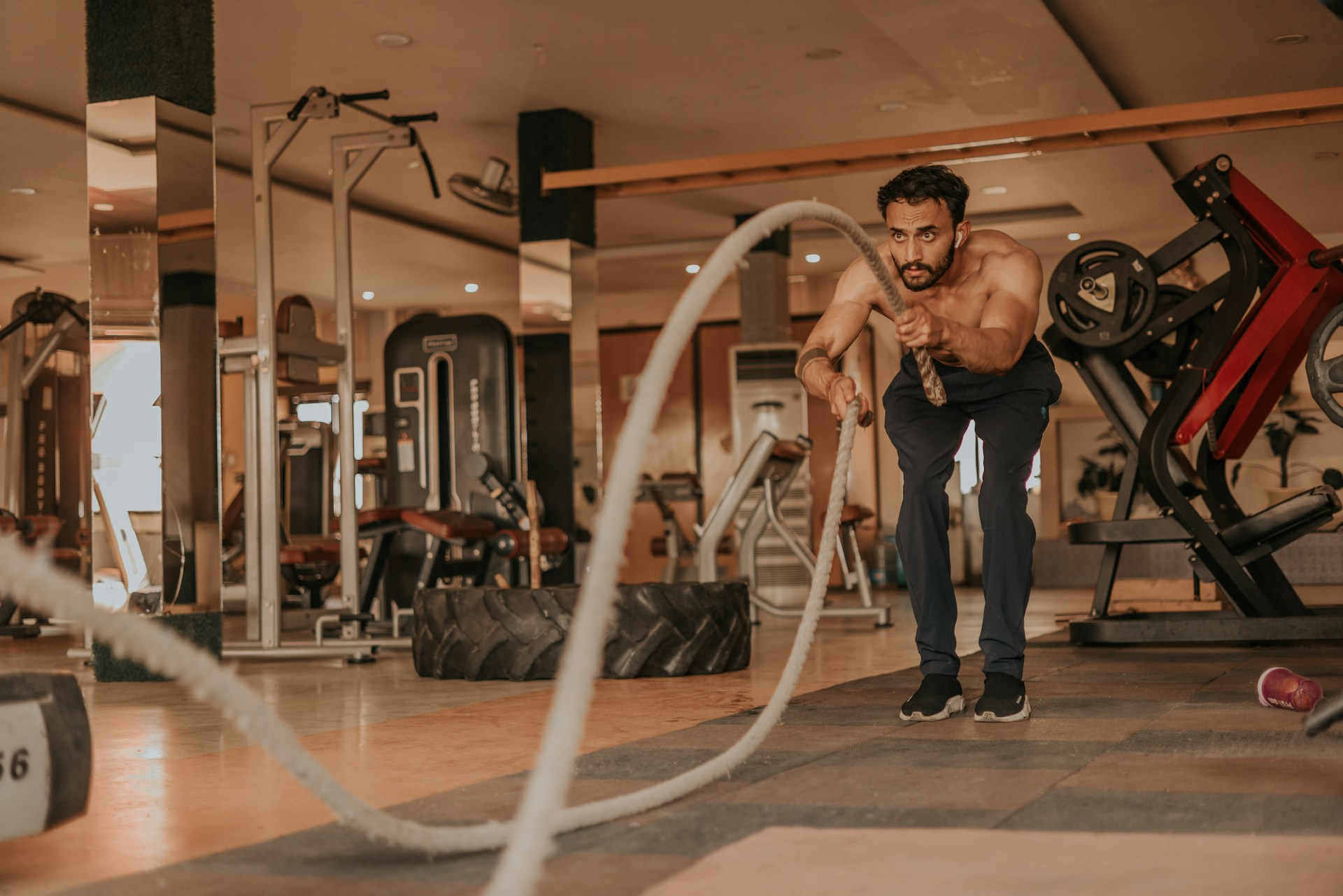 A shirtless man exercises with battle ropes in a spacious gym filled with various fitness equipment including weight machines, a large tire, and benches. He is in an athletic stance, gripping the ropes and generating waves across the floor. The environment is well-lit and modern, with mirrors and polished surfaces. The emotional tone is energetic and focused, suggesting intense physical training.