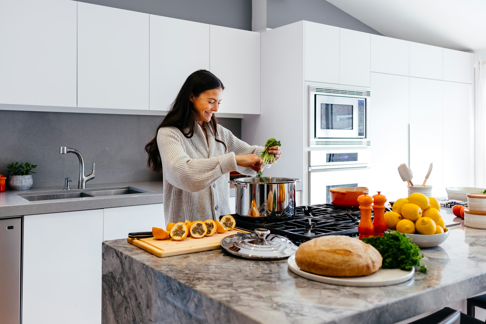 A person in a light sweater stands in a modern kitchen, preparing food by adding fresh greens to a large pot on the stove. The kitchen features white cabinets, a marble countertop with a loaf of bread, lemons, and cut passion fruit, and is brightly lit with a clean, organized atmosphere. The mood is focused and relaxed, suggesting home cooking or meal preparation.