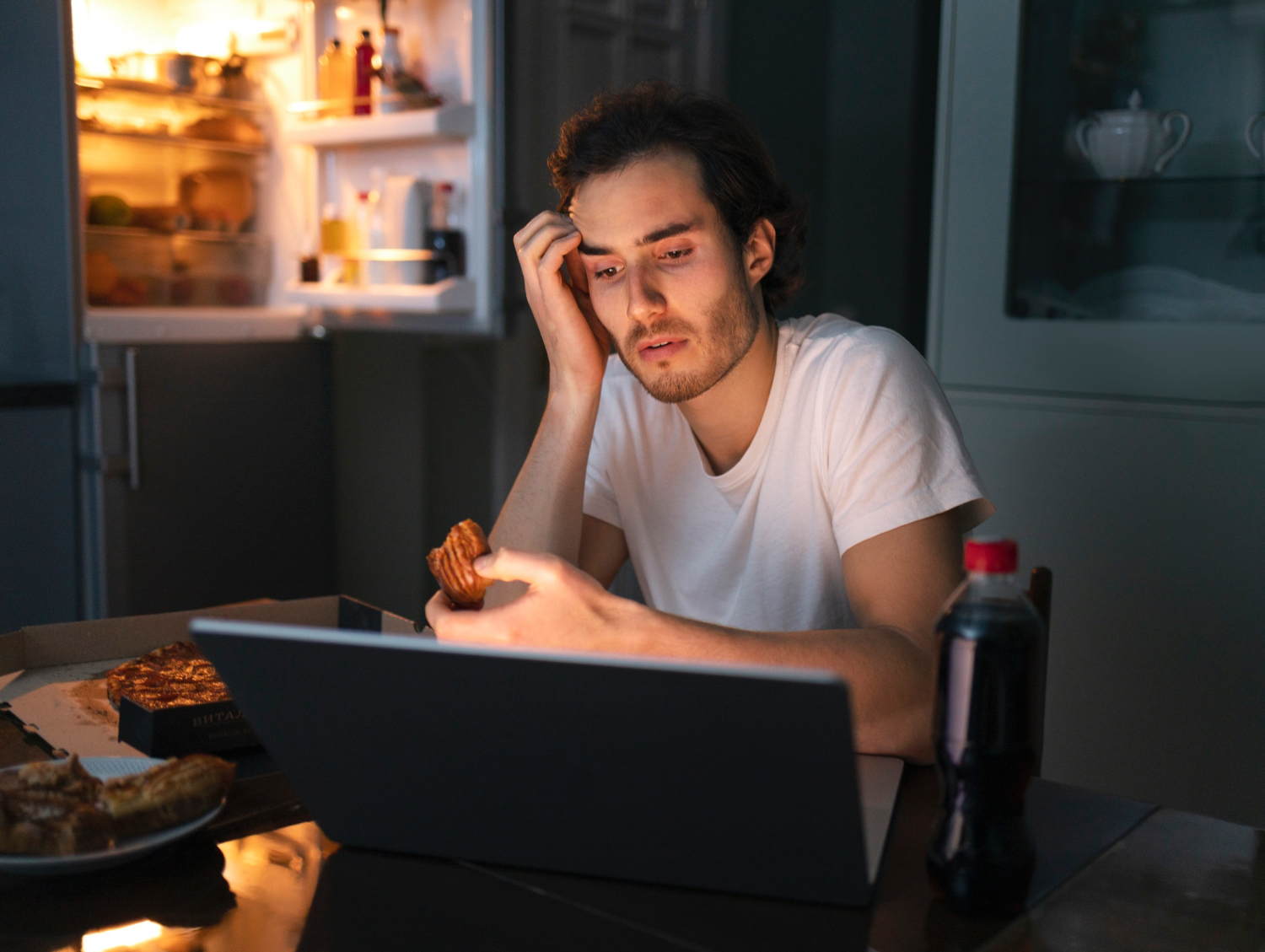 A person wearing a white t-shirt sits at a table in a dimly lit kitchen at night, eating a pastry while looking at a laptop screen. An open refrigerator with illuminated shelves is visible in the background, along with a pizza box, a plate of pastries, and a bottle of soda on the table. The environment feels quiet and solitary, suggesting late-night snacking or working.