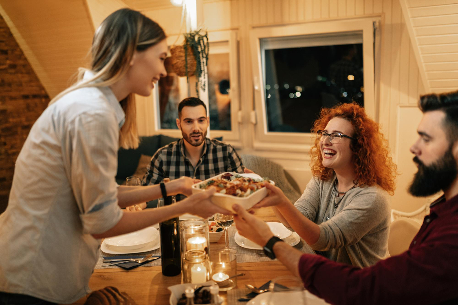 Friends gather around a warmly lit dinner table at night while one person serves food.