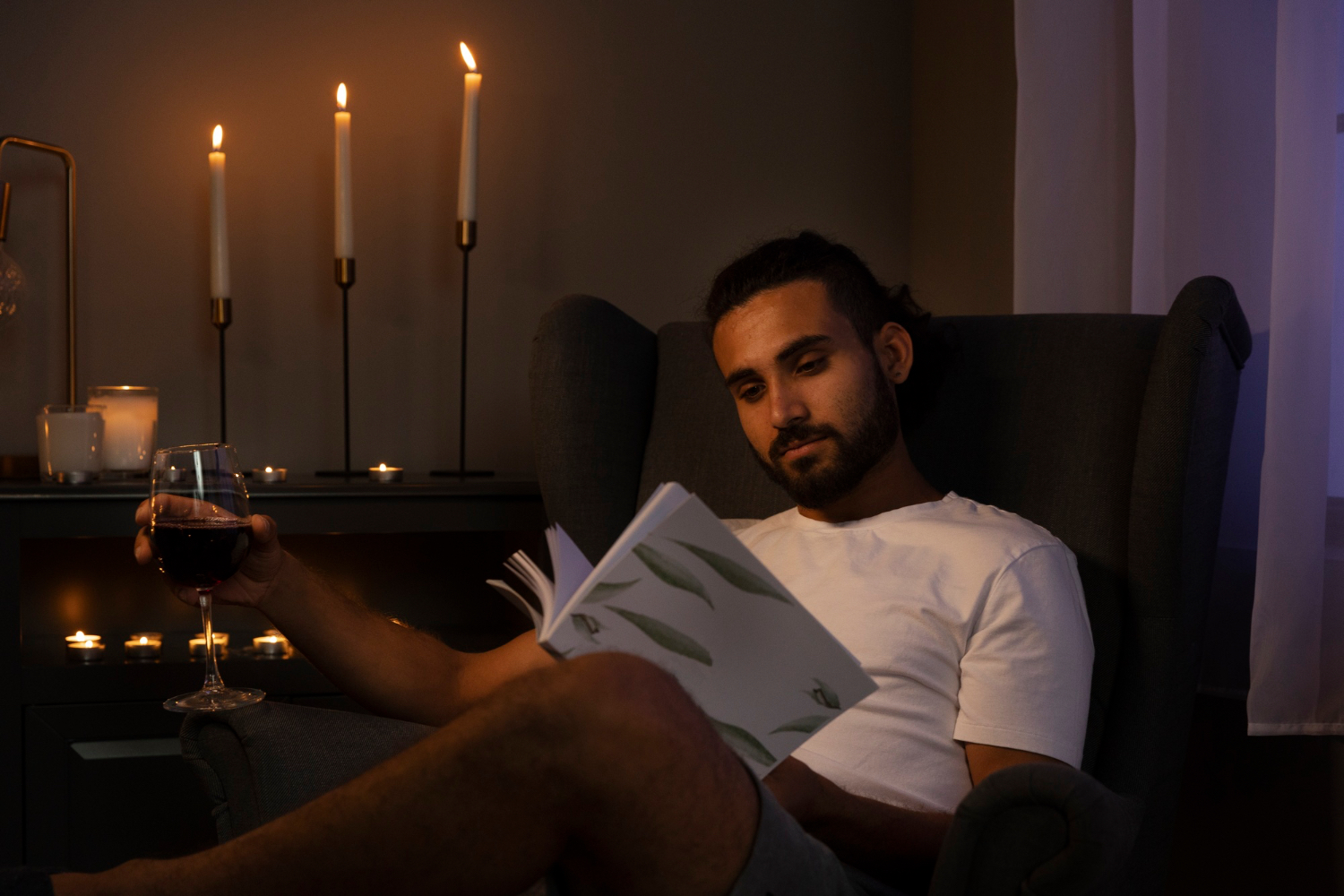 A man sits in a dark armchair reading a book by candlelight while holding a glass of red wine in a dim evening room.