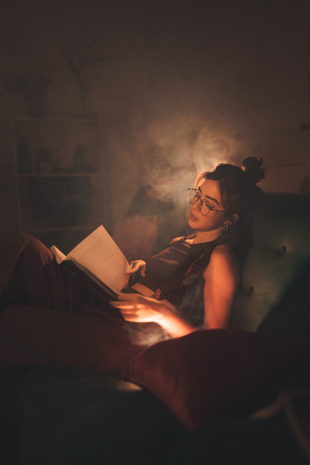 A woman reclines in a chair under soft amber light, reading with a sleepy expression in a quiet evening setting.