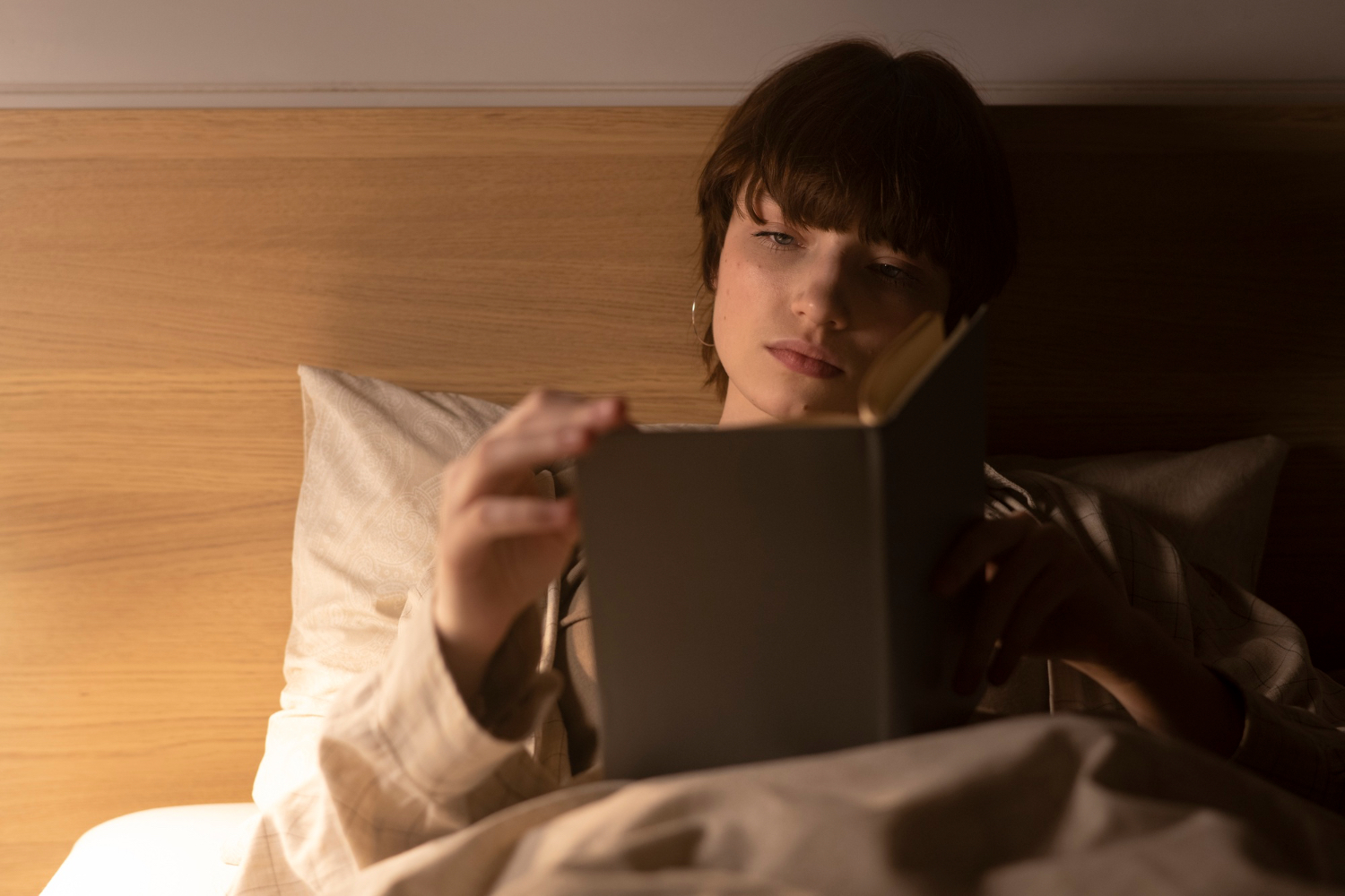 A person sits up in bed reading by a warm bedside light, with pillows and blankets around them in a calm nighttime setting.