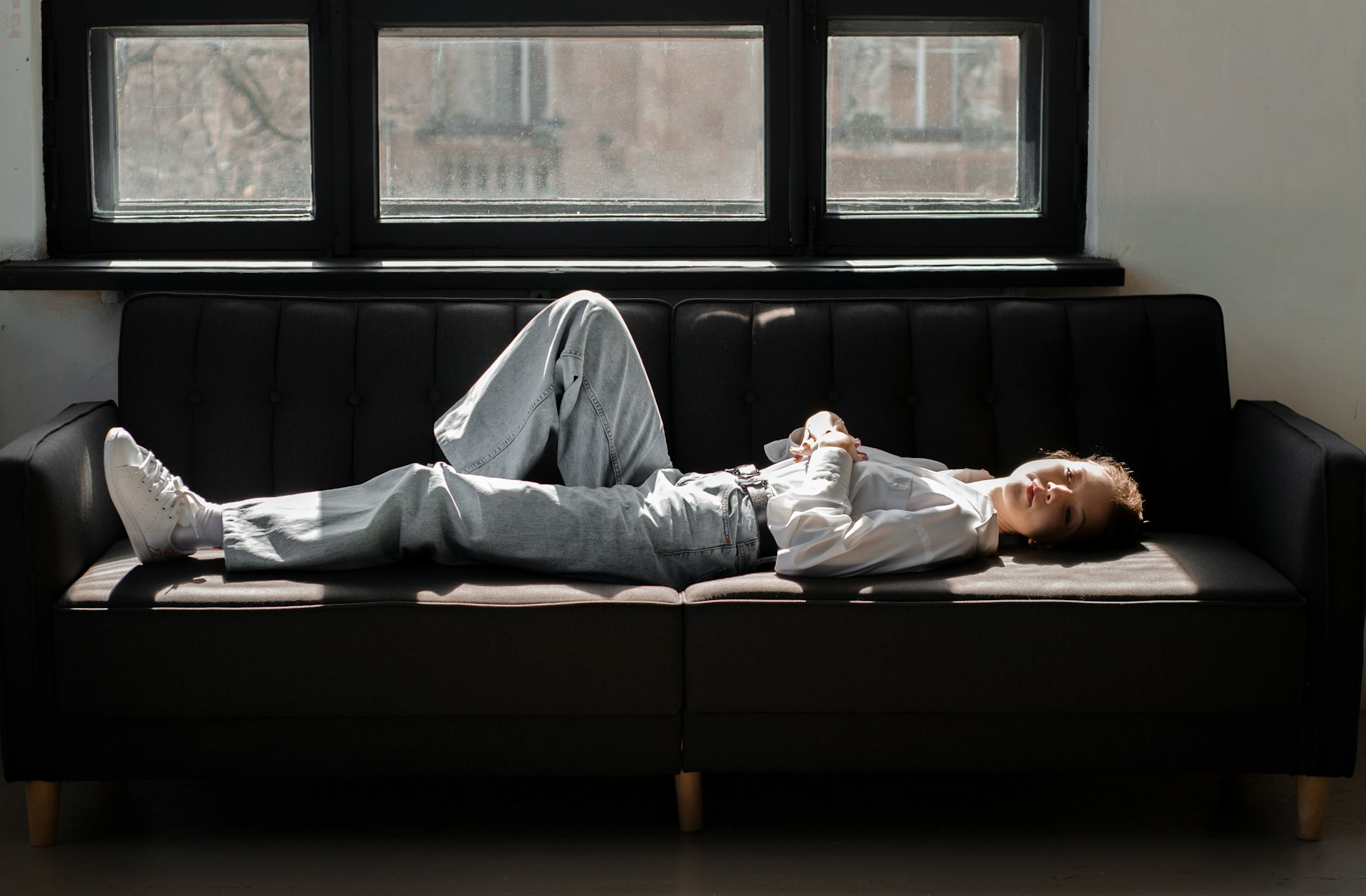 A person rests on a sofa in a sunlit living room, covered with a blanket and holding a book. The room includes a coffee table with a cup and a potted plant, and large windows letting in natural light. The environment feels cozy and peaceful, suggesting relaxation or napping during the day.