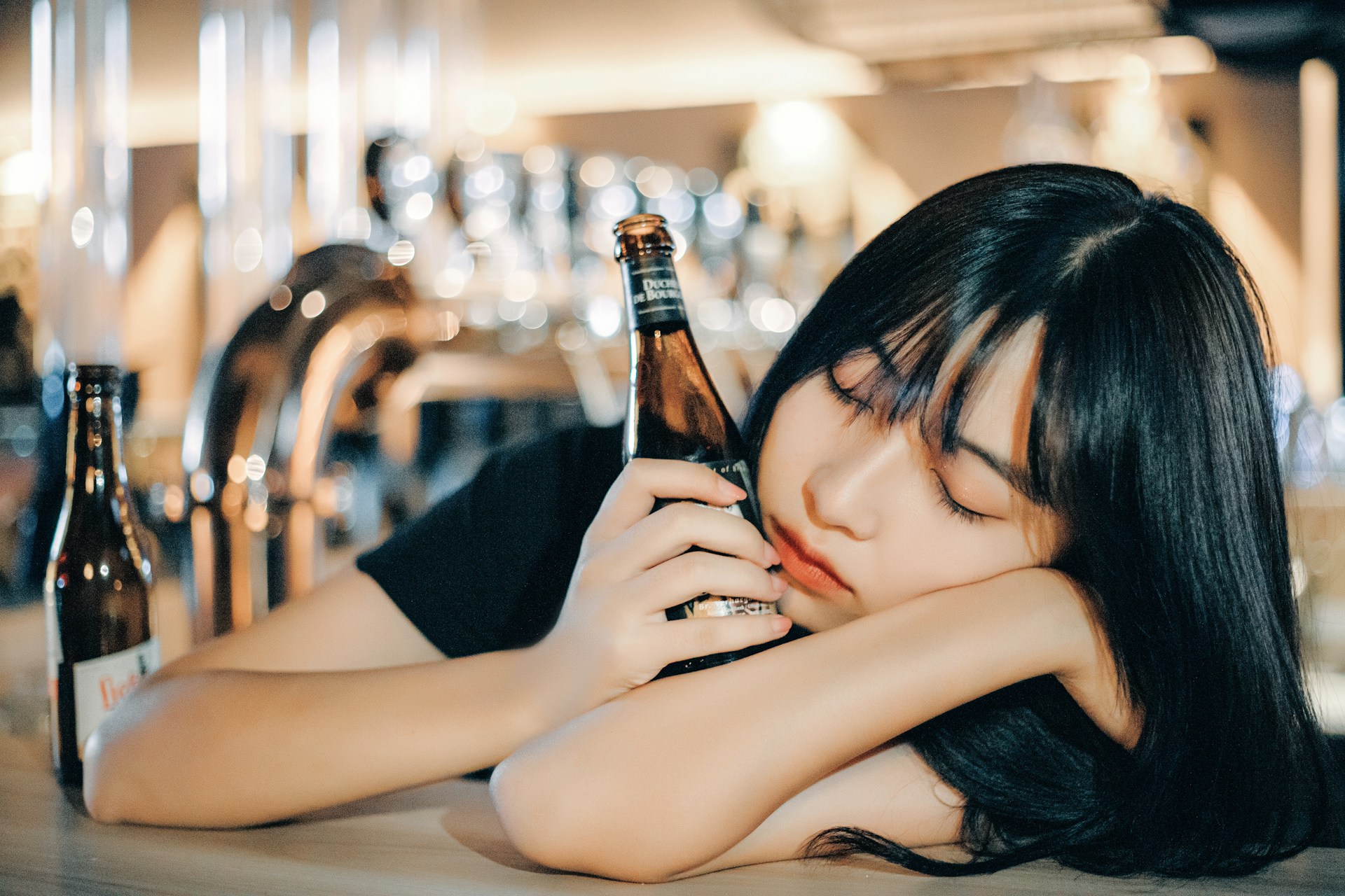 A person with long dark hair sits at a bar, resting their head on their arm while holding a brown beer bottle. Another beer bottle is on the counter nearby. The background features shiny metal taps and softly blurred lights, creating a relaxed and casual atmosphere.