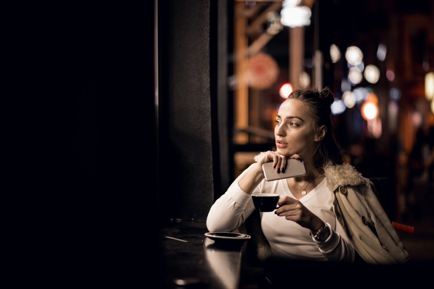 A woman sits at a café table at night, holding a smartphone in one hand and a cup of coffee in the other. She wears a light-colored top and a coat draped over her shoulders. The background features softly blurred city lights and a warm, intimate atmosphere, suggesting quiet reflection or relaxation.