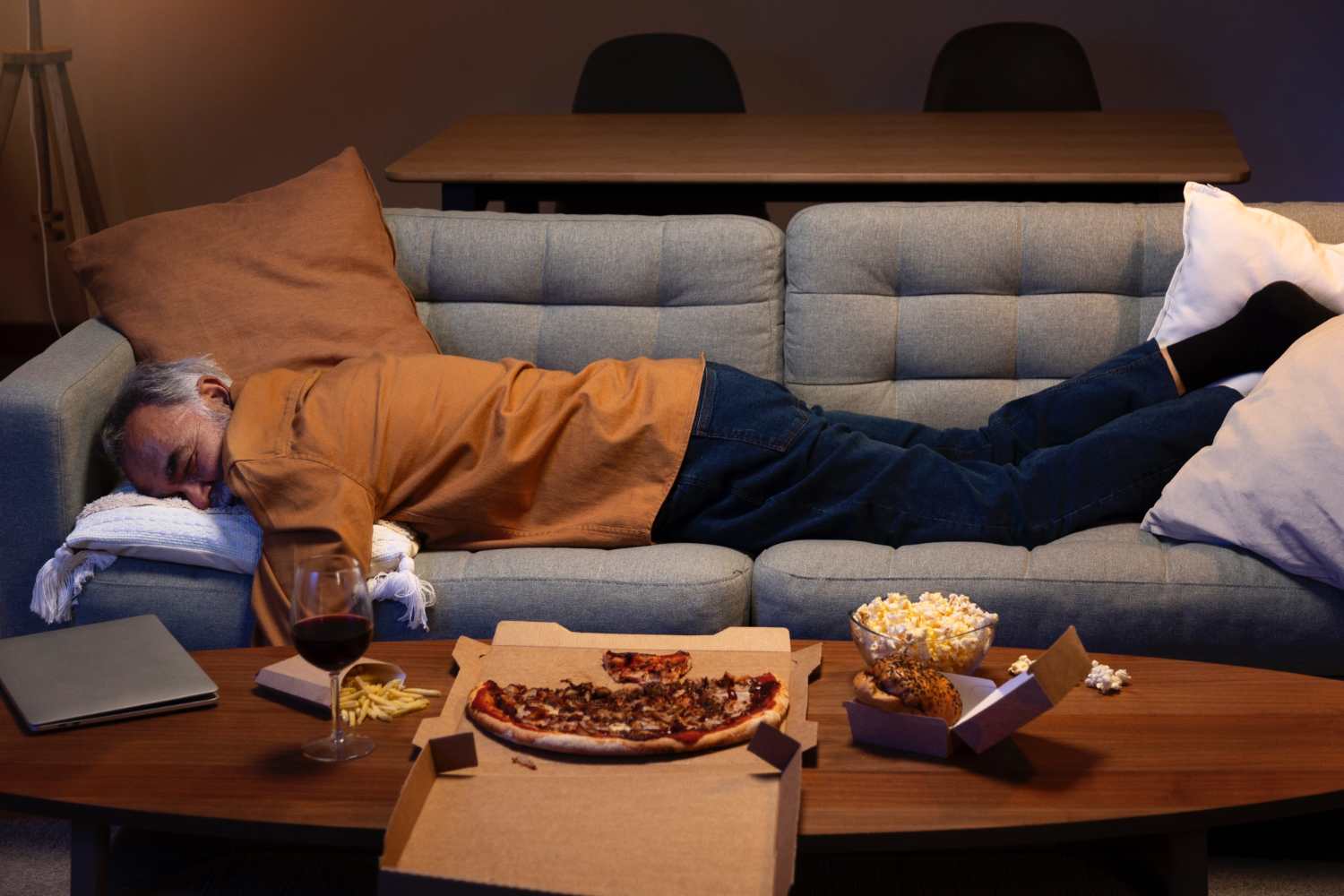 A person lies stretched out on a gray sofa in a living room, appearing to be asleep or resting. The coffee table in front of the sofa holds a half-eaten pizza in a box, a glass of red wine, a bowl of popcorn, a burger in a takeout container, and a few fries. A closed laptop sits nearby. The room is softly lit, with a dining table and chairs visible in the background. The atmosphere feels relaxed and casual, suggesting an evening of comfort and indulgence.