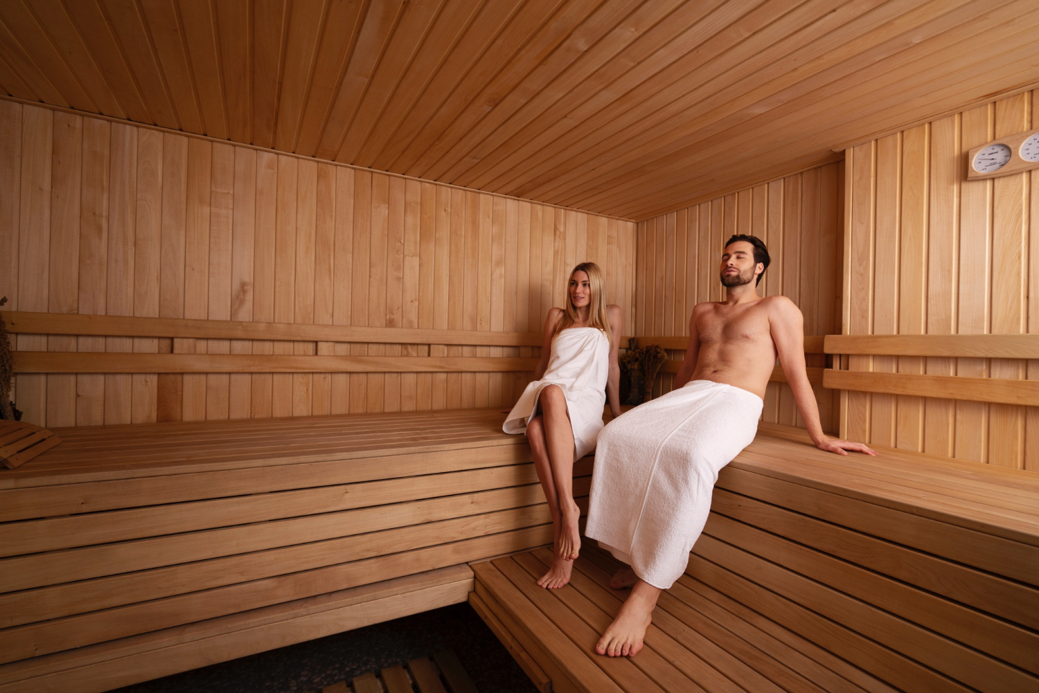 Two people wearing white towels sit and relax on wooden benches inside a spacious sauna lined with light-colored wood panels. The environment is warm and inviting, with a calm and tranquil atmosphere. A thermometer and hygrometer are mounted on the wall, indicating temperature and humidity levels.