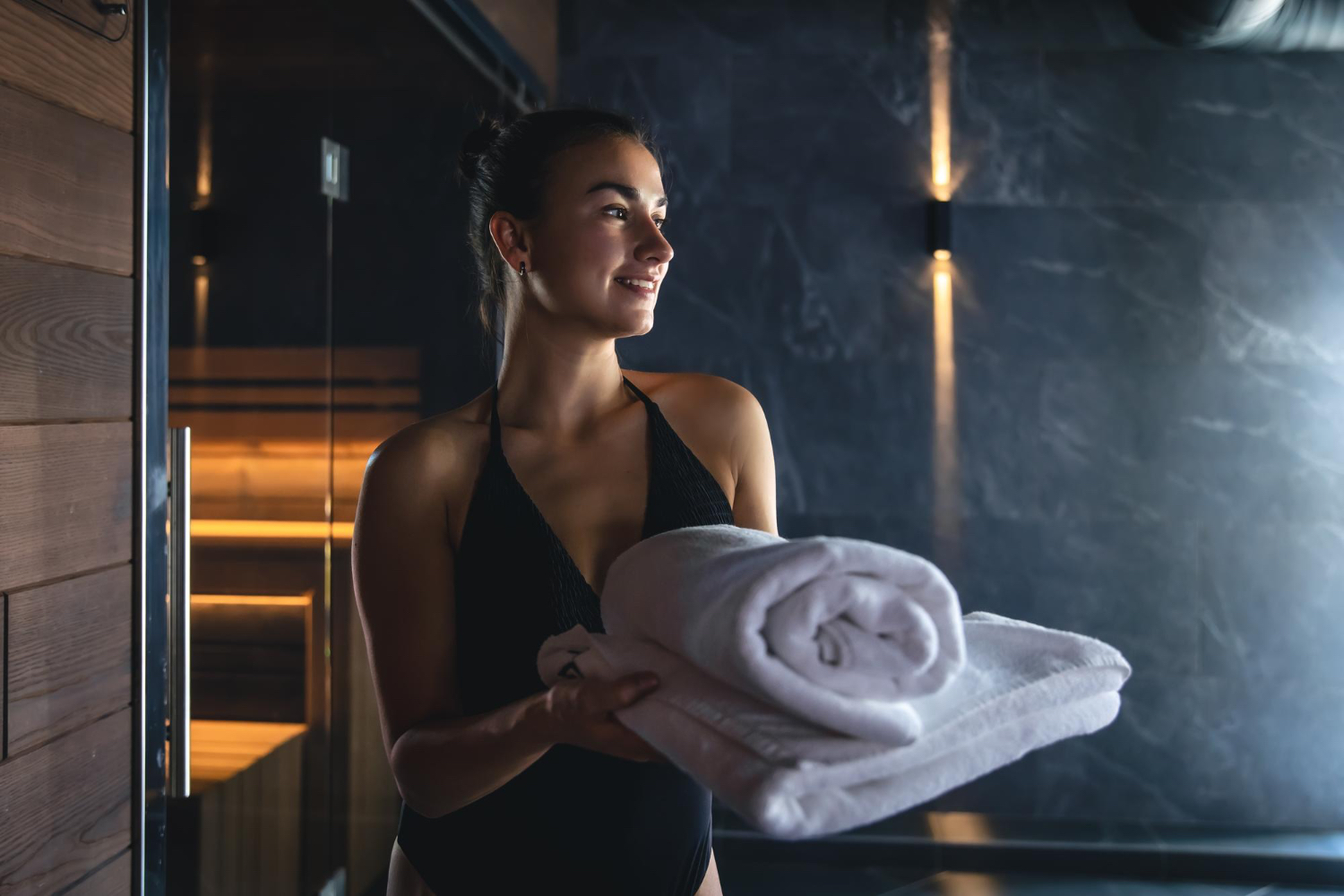 A woman wearing a black swimsuit stands indoors holding a stack of folded white towels in her arms. The background features a modern spa or sauna environment with dark walls, warm lighting, and wooden accents. The atmosphere feels calm, inviting, and tranquil, suggesting relaxation or preparation for a spa experience.