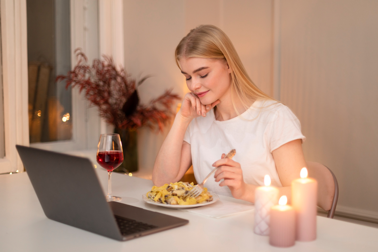 A woman eating a pasta dinner at night beside a laptop and candlelight.