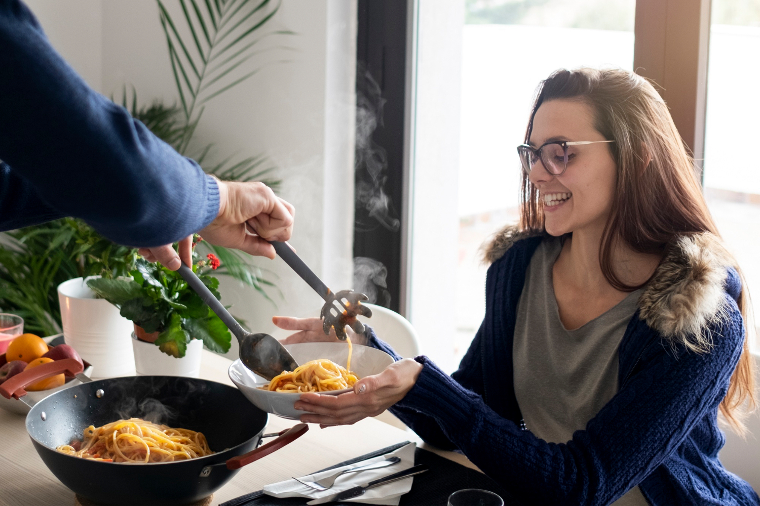 A woman is served a warm bowl of pasta at an indoor dinner table.