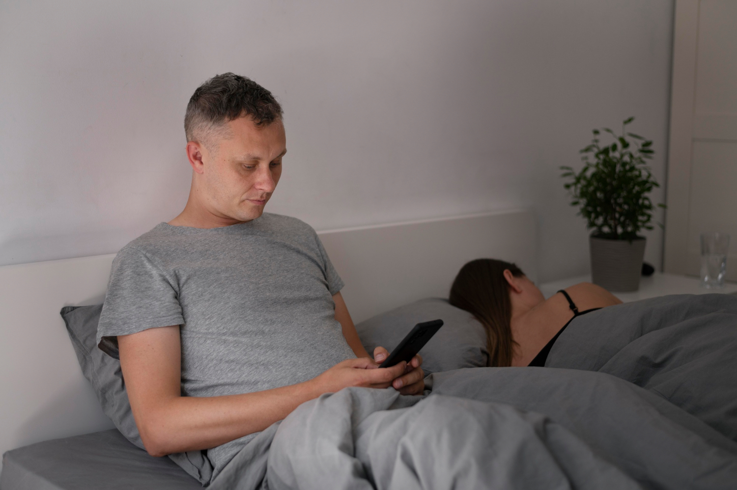 A man sits awake in bed looking at his phone while another person sleeps beside him, illustrating how screen use and poor wind-down habits can keep rest-day sleep off track.