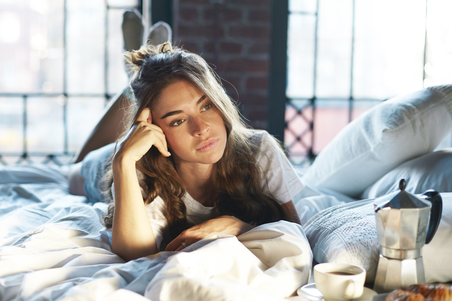 A woman lies awake in bed beside a cup of coffee and a moka pot, looking tired in the morning light.