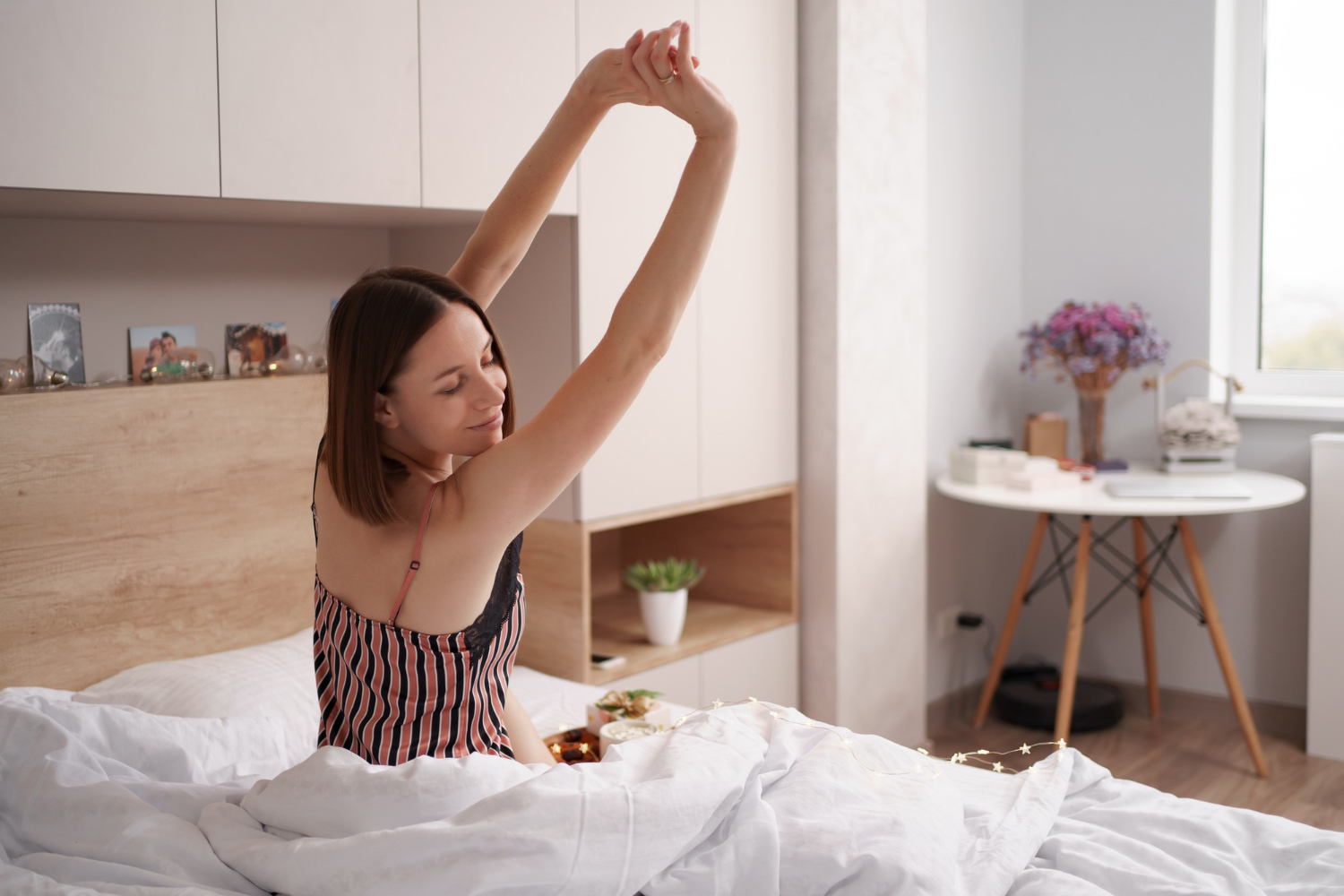 A woman sits up in bed and stretches her arms overhead in a bright, calm bedroom.